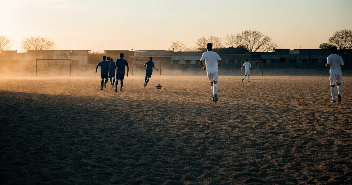 Ein sandiger Fußballplatz am Rand einer Stadt in warmem Abendlicht, Staub in der Luft und zwei Tore in der Ferne.