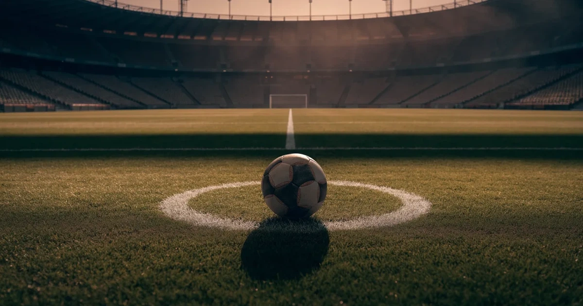 Ein Fußball liegt auf dem Anstoßpunkt eines leeren Rasenplatzes, im Hintergrund eine große Stadiontribüne im Abendlicht.
