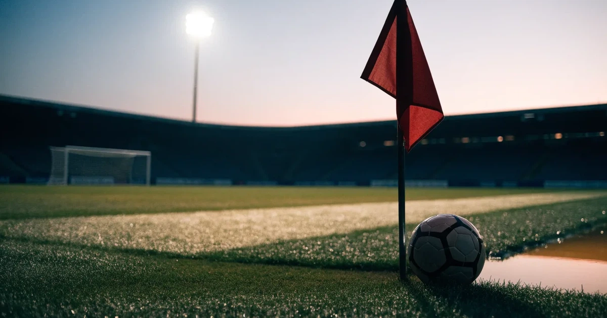 Blick durch ein Stadiontor auf einen grünen Rasen im Abendlicht, ein Ball ruht an der Eckfahne, die Tribünen sind leer.