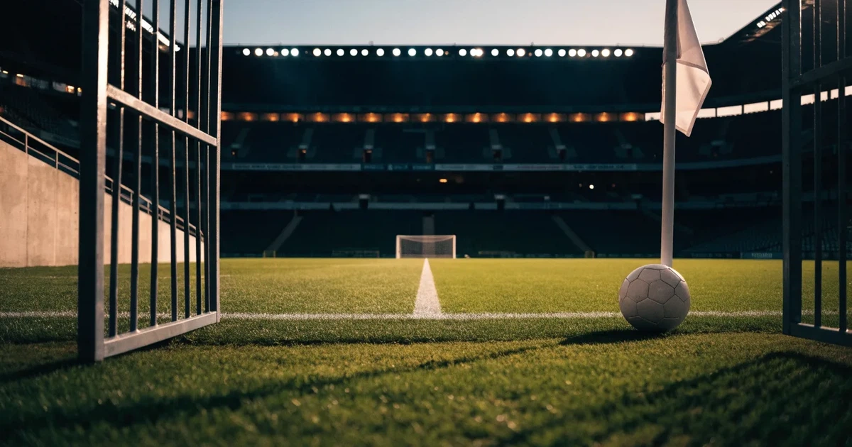 Blick durch ein Stadiontor auf einen grünen Rasen im Abendlicht, ein Ball liegt an der Eckfahne, die Ränge sind leer.