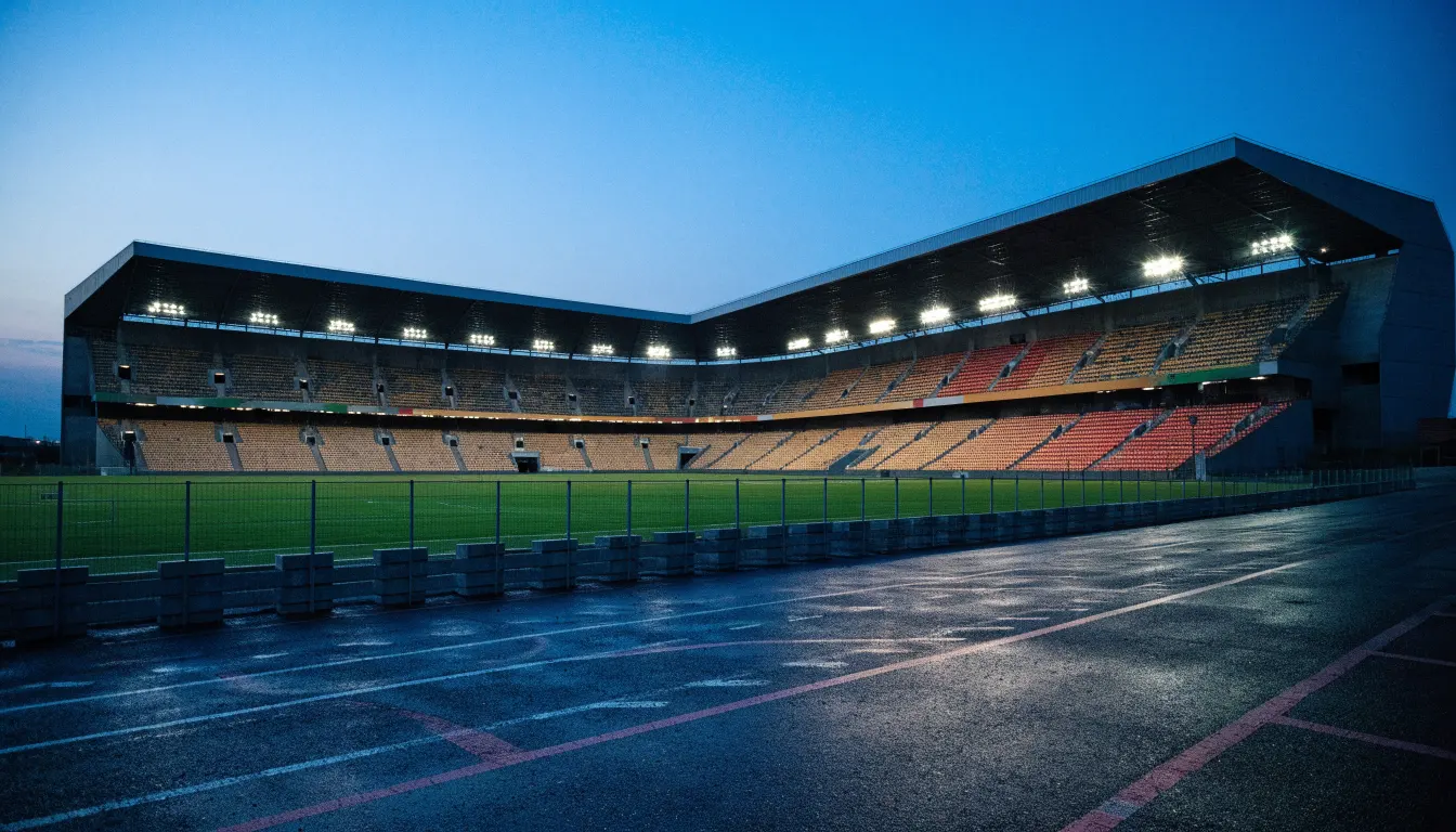 Ein Stadion unter Flutlicht aus der Ferne gesehen, davor ein leerer Parkplatz in der Dämmerung.