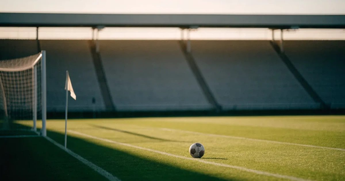 Ein Fußballplatz mit frisch gemähtem Rasen im Abendlicht, ein Ball liegt im Strafraum, im Hintergrund unscharf eine leere Tribüne.
