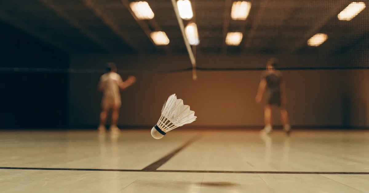 Eine leere Badmintonhalle mit gespanntem Netz und hellen Hallenlichtern, ein Federball liegt auf dem Hallenboden.