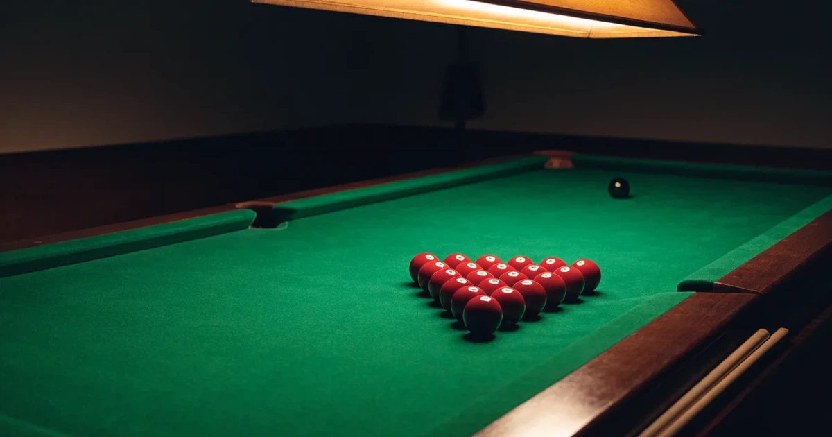 Ein Snookertisch mit grünem Tuch und aufgebauten roten Kugeln in einem abgedunkelten Raum unter einer hängenden Lampe.
