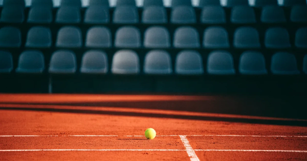 Ein Sandplatz mit rötlichem Belag und gezogenen weißen Linien, ein einzelner Tennisball liegt neben der Linie, im Hintergrund eine leere Tribüne.