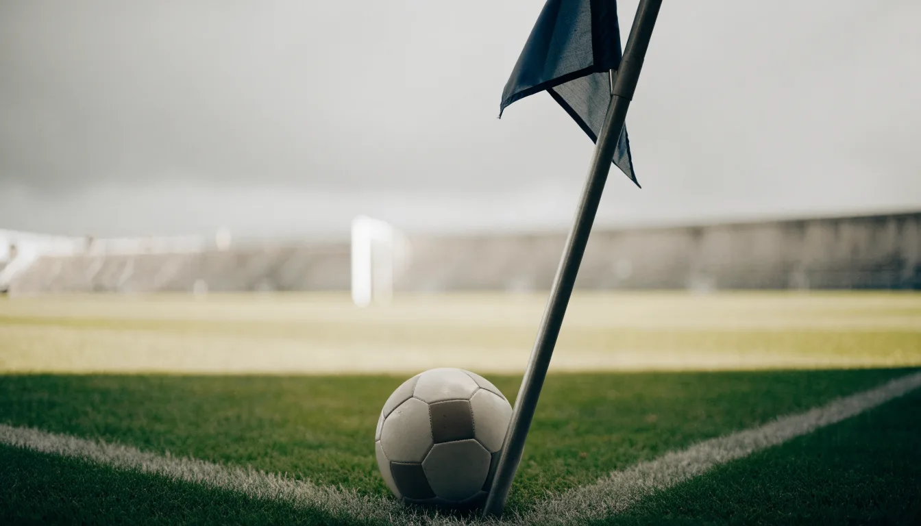 Ein Fußball liegt neben einer Eckfahne auf kurz gemähtem Rasen unter bewölktem Himmel.