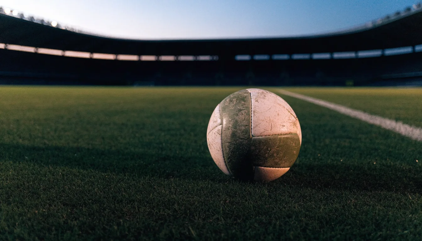 Ein Fußball liegt auf einem Rasenplatz in der Dämmerung, im Hintergrund ein ferner Stadionrand unter Flutlicht.